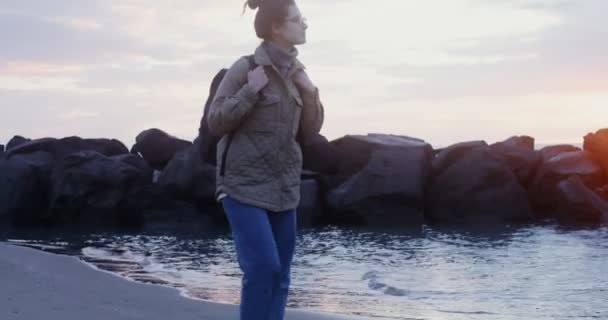 Mladá, vřele oblečená žena kráčí po pobřeží při západu slunce — Stock video