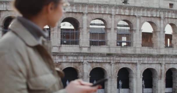 Egy nő tárcsáz egy üzenetet a mobilján, miközben a Colosseum előtt áll. — Stock videók