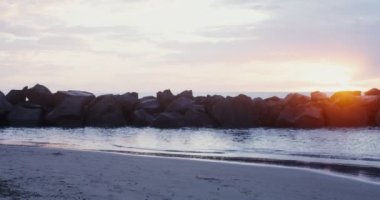 Sıcak giyimli genç bir kadın gün batımında deniz kıyısında yürüyor.