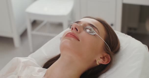 Uma paciente mulher em óculos de proteção está em uma sala de cosmetologia — Vídeo de Stock