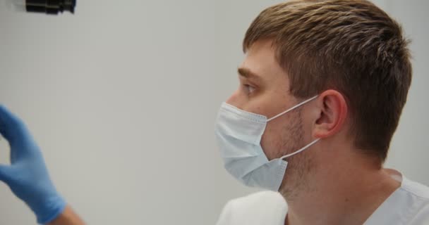 Un médico o científico masculino con una máscara médica mira a través de un microscopio — Vídeo de stock
