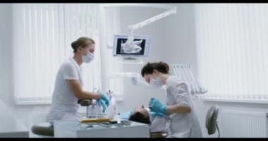 Bir kadın diş hekimi, dişçi koltuğunda yatan bir hastanın ağız boşluğunu muayene eder.