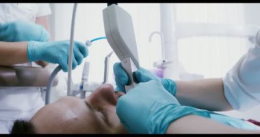 Bir hasta dişçi koltuğunda yatarken doktor dişlerini kamerayla muayene ediyor.