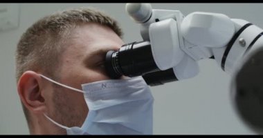 Tıbbi maskeli bir erkek doktor ya da bilim adamı mikroskopla bakar.