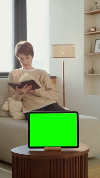 Μια γυναίκα που διαβάζει ένα βιβλίο κάθεται σε έναν καναπέ μπροστά από ένα πράσινο tablet οθόνη — Αρχείο Βίντεο