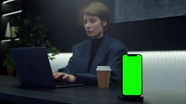 Un cellulare con schermo verde si trova sul desktop di fronte alla donna che lavora al computer portatile — Video Stock