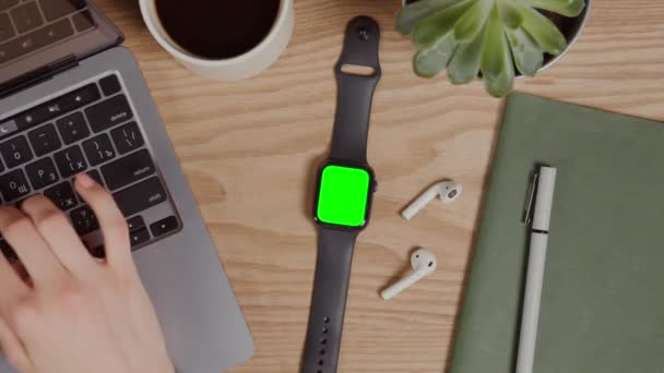 Relógio digital com uma tela verde deitada na mesa ao lado de mãos digitando no laptop — Vídeo de Stock