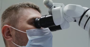 Tıbbi maskeli bir erkek doktor ya da bilim adamı mikroskopla bakar.