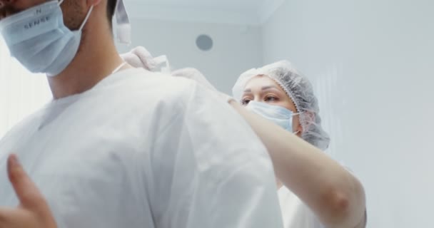 Hemşire, bir dişçiye beyaz steril bir elbise giydirip, sırtının bağını bağlamasına yardım eder. — Stok video