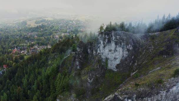 Un villaggio tra una foresta ai piedi di una rupe rocciosa ricoperta di nuvole — Video Stock
