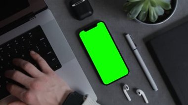 Yeşil ekranlı bir cep telefonu dizüstü bilgisayarda yazan bir adamın masasında duruyor.