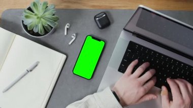 Yeşil ekranlı bir cep telefonu dizüstü bilgisayarda yazan bir adamın masasında duruyor.