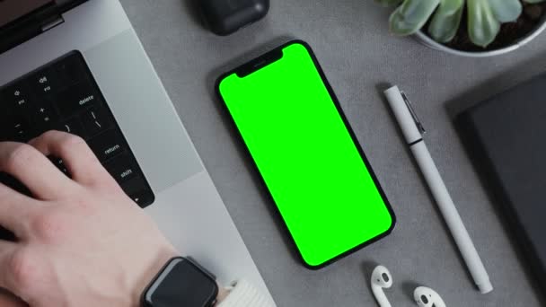 Un teléfono móvil con una pantalla verde se encuentra en el escritorio mans escribiendo en un ordenador portátil Video de stock