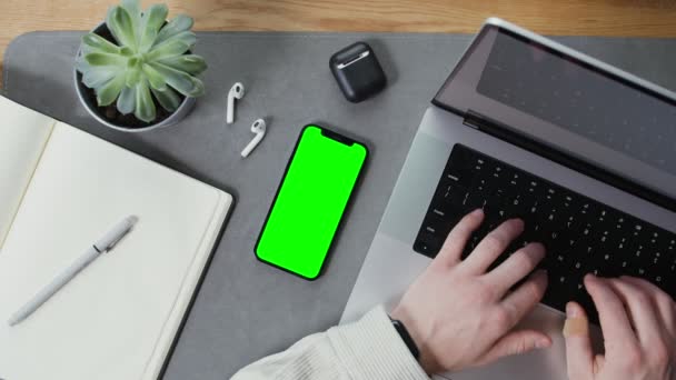 Ένα κινητό τηλέφωνο με πράσινη οθόνη βρίσκεται στην επιφάνεια εργασίας mans πληκτρολόγηση σε ένα φορητό υπολογιστή — Αρχείο Βίντεο