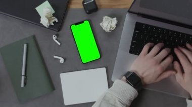 Bir adam yanında cep telefonu olan bir masaüstünde dizüstü bilgisayar kullanıyor.