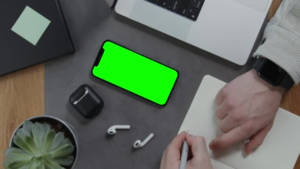 Ein Mann macht sich Notizen in einem Notizbuch, das neben einem Mobiltelefon mit grünem Bildschirm liegt Videoclip