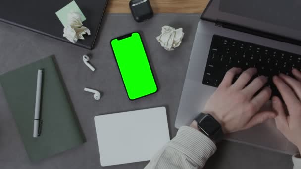 Egy férfi gépel egy laptopon egy asztalnál, mellette egy mobiltelefonnal. — Stock videók