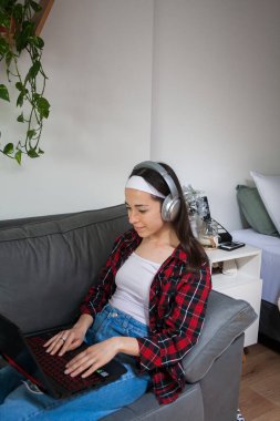 Evde dizüstü bilgisayarla çalışan ve koltukta oturmuş kulaklıkla müzik dinleyen genç bir kadın..