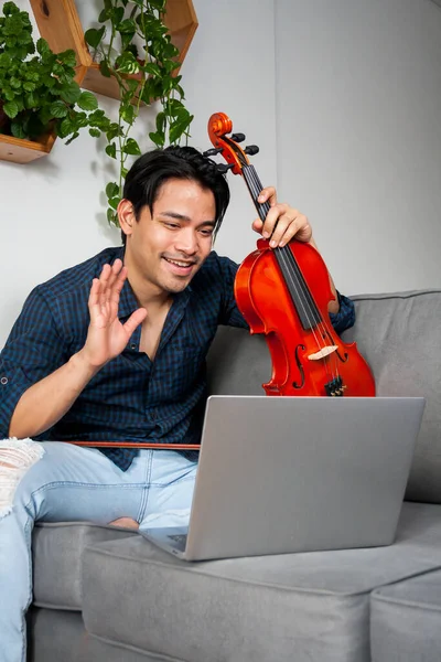 Young Hispanic man, music student making a video call with a laptop, showing off his musical instrument.
