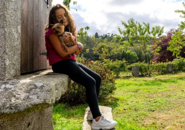 Taş bir inşaatta sopayla dövülen uzun saçlı kız kucağında küçük bir köpek tutuyor.