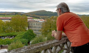 Arka planda binaların ve ormanların görüldüğü terasa bakan turuncu tişörtlü orta yaşlı bir adam.