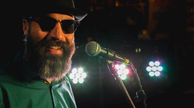 Mikrofonun önündeki sakallı adam. Arka planda renkli ışıklar var.