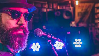 Mikrofonun önündeki sakallı adam. Arka planda renkli ışıklar var.