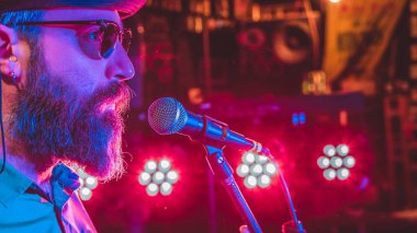 Mikrofonun önündeki sakallı adam. Arka planda renkli ışıklar var.