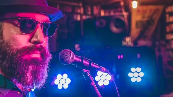 Mikrofonun önündeki sakallı adam. Arka planda renkli ışıklar var.