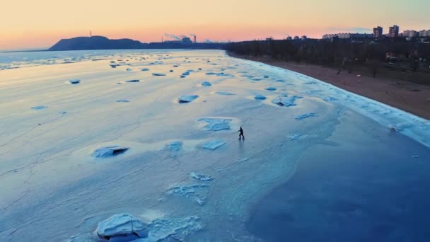 Ein Mann läuft bei Sonnenuntergang auf dem Eis des gefrorenen Meeres nahe der Küste der Industriestadt Schlittschuh. Eislauftraining. Winterfreizeit am Meer. Drohnenaufnahmen. — Stockvideo