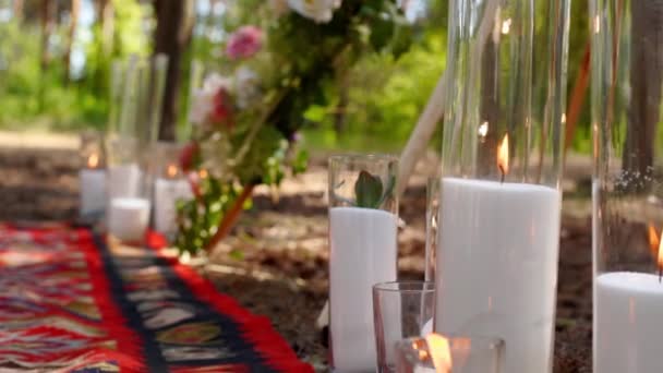 Свечи, горящие в стеклянных вазах, фляжки на ковре возле богемной арки типи, украшенные розами и цветами, завернутыми в сказочные огни на открытой свадебной церемонии в сосновом лесу. Бохо деревенский декор. — стоковое видео