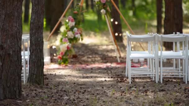 Schöne böhmische Tipi-Bogendekoration am Ort der Trauung im Freien im Kiefernwald mit Zapfen. Stühle, floristische Blumenkompositionen aus Rosen, Teppich, Lichterketten. Ländliche Sommerhochzeit. — Stockvideo