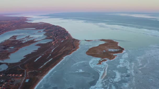 Légi kilátás a kis tengerparti város a fagyos téli naplementében. Tengerpart szigettel, tavakkal és öblökkel. A település a fagyott tenger közelében. Téli tengerparti falu éjszakai fényei. — Stock videók