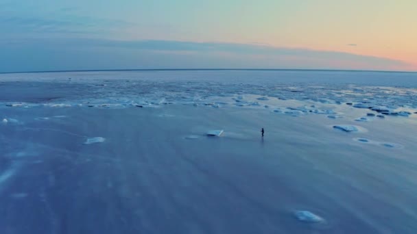 Een man is schaatsen op het ijs van de bevroren zee in de buurt van de kust van de stad bij zonsondergang. Schaatstraining. Wintersport aan zee. Drone-beelden. — Stockvideo