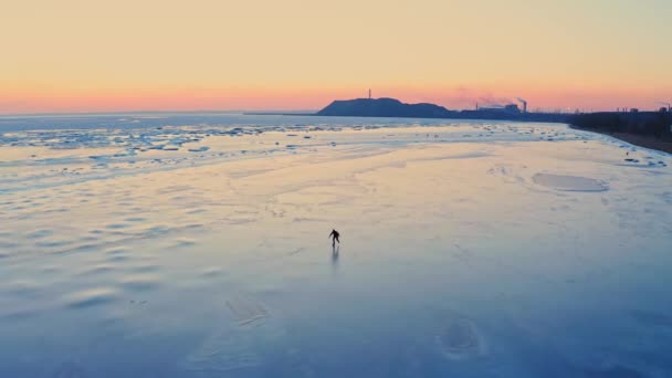 Чоловік катається на льоду замерзлого моря біля узбережжя промислового міста на заході сонця. Тренування з катання на ковзанах. Зимове дозвілля на узбережжі. Мультфільми дронів . — стокове відео