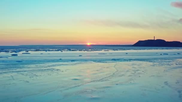 氷結した冬の海の上の日没の空中ビュー。無人機は日の出に海の上に降下する。融解氷のドリフトと産業都市の上空を飛行する。地球温暖化。鮮やかなカラフルな空。工業プラント. — ストック動画