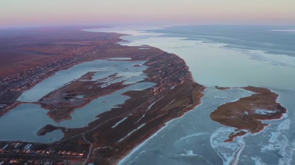 Letecký pohled na malé přímořské městečko při mrazivém zimním západu slunce. Mořské pobřeží s ostrovem, jezery a zálivy. Osídlení u zamrzlého moře. Noční světla vesničky na pobřeží. — Stock video