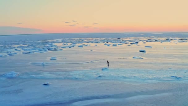 男は日没時に町の海岸近くの凍結した海の氷の上でスケートをしている。アイススケートのトレーニング。海辺で冬のレジャー。ドローン映像. — ストック動画