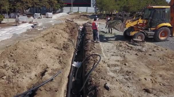 Ukraine, Mariupol - September 1, 2020. Workers digging trench on construction site. Loader tractor pours gravel of bucket. Underground channel with electric power cables. Infrastructure building. — Stok video