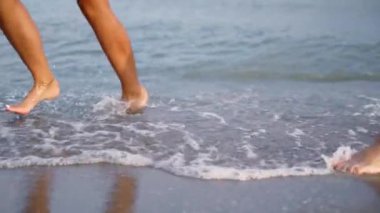 A couple is running, dancing along the surf line. Happy young people have fun on the sandy beach in summer. Slender tanned legs in the sea waves