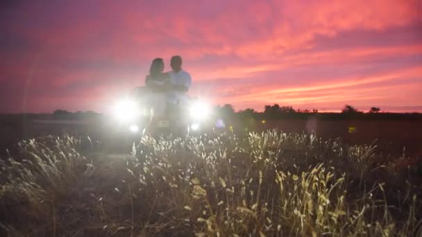 Een man en een vrouw zoenen bij de auto in het veld. De koplampen van de auto verlichten het silhouet van een verliefd stel. Een zachte familieknuffel bij zonsondergang. Familie reis met de auto over de steppe. — Stockvideo