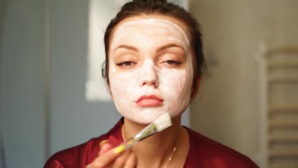 女性はブラシで顔にクレンジングマスクを適用します。女性は顔のスキンケア手順を行います.女の子はクリームで肌をこすり、保湿 . — ストック動画