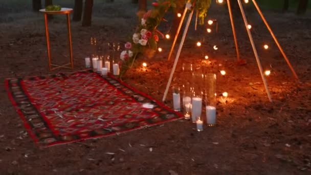 Bohemisk tipi träbåge dekorerad med brinnande ljus, rosor och pampass gräs, insvept i älva ljus belysning på utomhus bröllop plats i tallskog på natten. Lökar körtel lyser. — Stockvideo