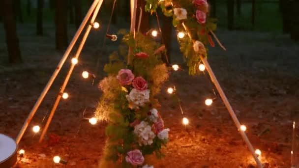 Boheemse tipi houten boog versierd met brandende kaarsen, rozen en pampass gras, verpakt in sprookjes verlichting op outdoor bruiloft locatie in dennenbos 's nachts. Bollen bloemenslinger schijnt. — Stockvideo