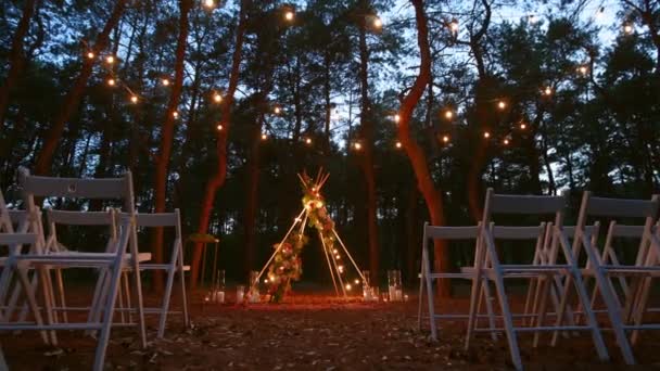 Świąteczne oświetlenie struny na boho tipi łuku wystrój na zewnątrz miejsce ceremonii ślubnej w sosnowym lesie w nocy. Stare lampki strunowe girlandy świecące nad krzesłami na letnim weselu wiejskim. — Wideo stockowe