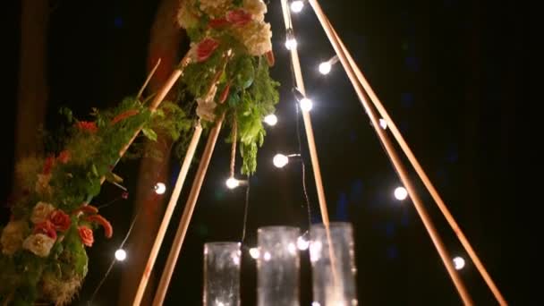 Bohemisk tipi träbåge dekorerad med brinnande ljus, rosor och pampass gräs, insvept i älva ljus belysning på utomhus bröllop plats i tallskog på natten. Lökar körtel lyser. — Stockvideo