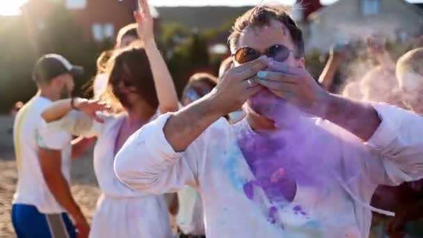 Handsome man claps hands with colored powder in and paints his moustache with lavender-blue dye at Holi festival. People celebrate hindu holiday. End of lockdown, covid pandemic, restrictions. — Stock Video