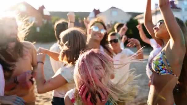 Cheerful girl celebrates Holi festival with friends stained in colored powder. People have fun at hindu holiday. Guys dance, jump on beach in slow motion. End of covid pandemic isolation — Stock Video