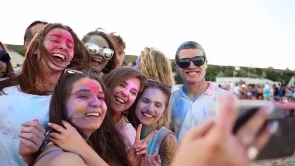Pessoas alegres cobertas de pó colorido levam selfie no smartphone e sorriem no festival Holi na praia em câmera lenta. Os amigos untados em cores secas tomam a foto de grupo pelo telefone. Fim da pandemia vívida — Vídeo de Stock