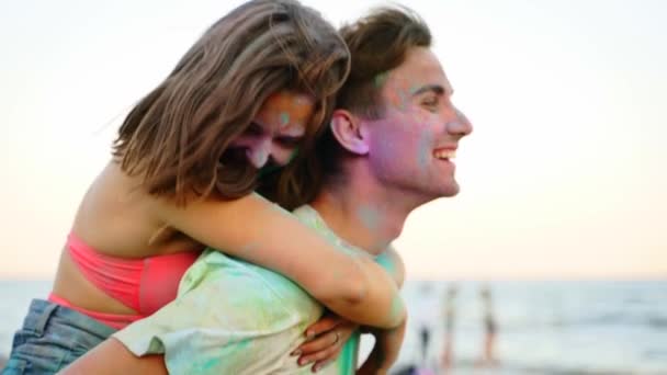Un homme souriant porte sur le dos une jolie femme tachée de poudre colorée au festival Holi sur la plage. Couple s'amuse avec des amis à hindou vacances de couleurs, l'amour, l'unité. Fin de l'isolement pandémique covide — Video
